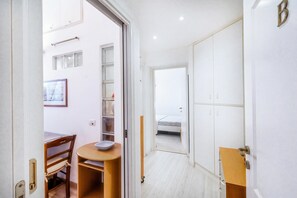 A narrow hallway with a wooden table and chair, a small shelf, and a door leading to a bedroom with a bed.