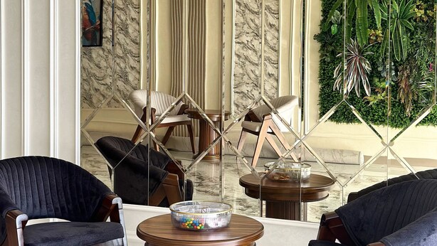 A modern lobby with a marble floor, dark upholstered chairs, a wooden round table, and a decorative glass partition. - Colibri Hotel  (Bukhara)