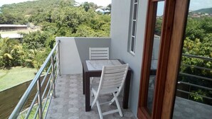Un balcón con mesa y silla, vistas a zonas verdes y edificios, y una barandilla de metal.