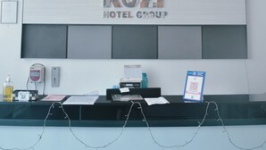 Un comptoir d’accueil avec une enseigne indiquant « KOTM HOTEL GROUP », un téléphone, un distributeur de gel hydroalcoolique et un code QR pour l’enregistrement.