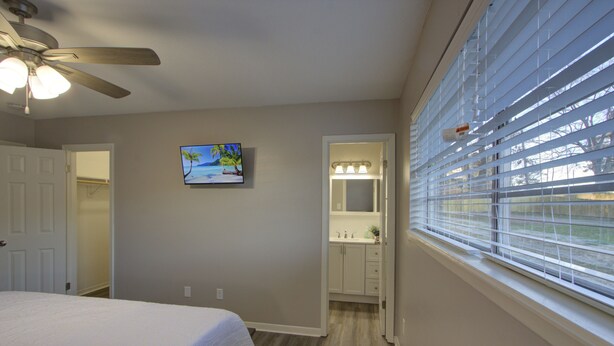 A bedroom with a bed, a ceiling fan, a TV on the wall, a bathroom with a mirror and sink, and a window with blinds. - A Quiet Place - Dogwood Retreat in Texarkana (Texarkana)