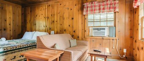 A cozy cabin room with a bed, a sofa, a wooden table, and a window with plaid curtains.