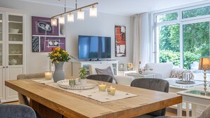 A modern living room with a wooden dining table, a TV, and a large window with a view of greenery.