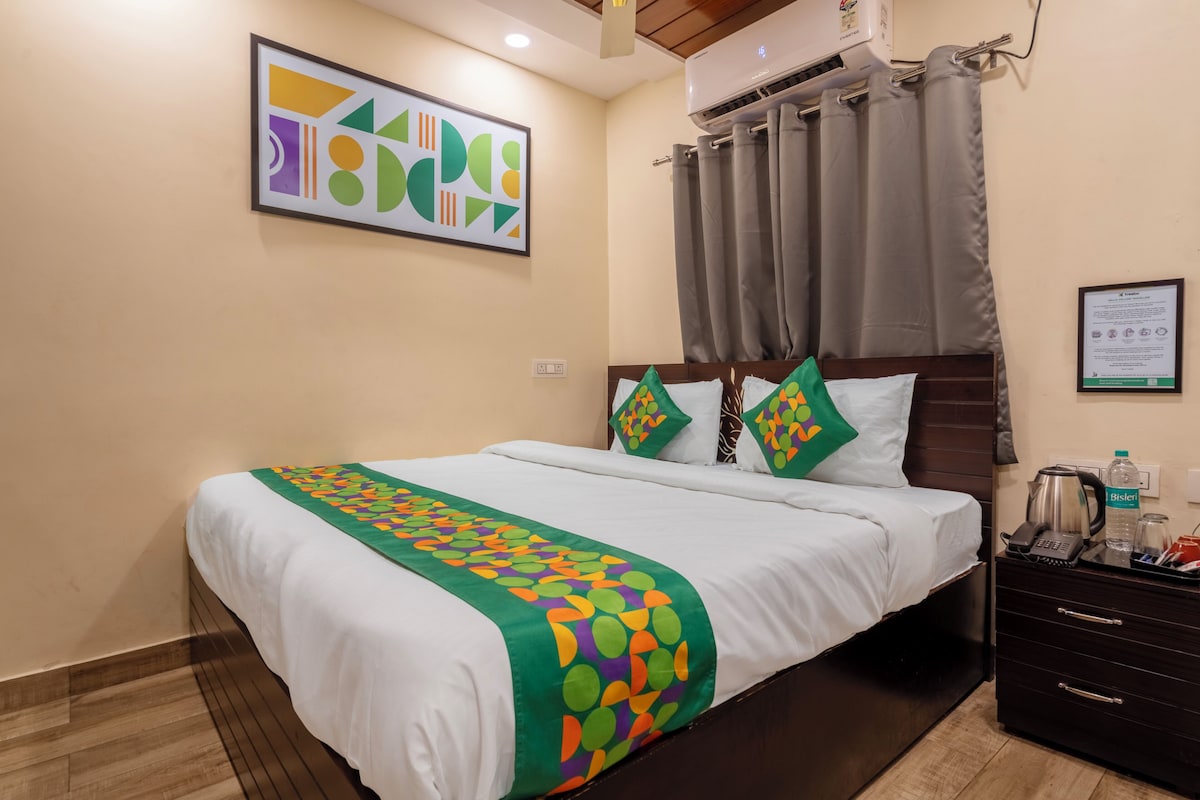 A hotel room with a bed, a wall-mounted air conditioner, a framed abstract artwork, a grey curtain, a bedside table with a kettle, and a framed certificate on the wall.