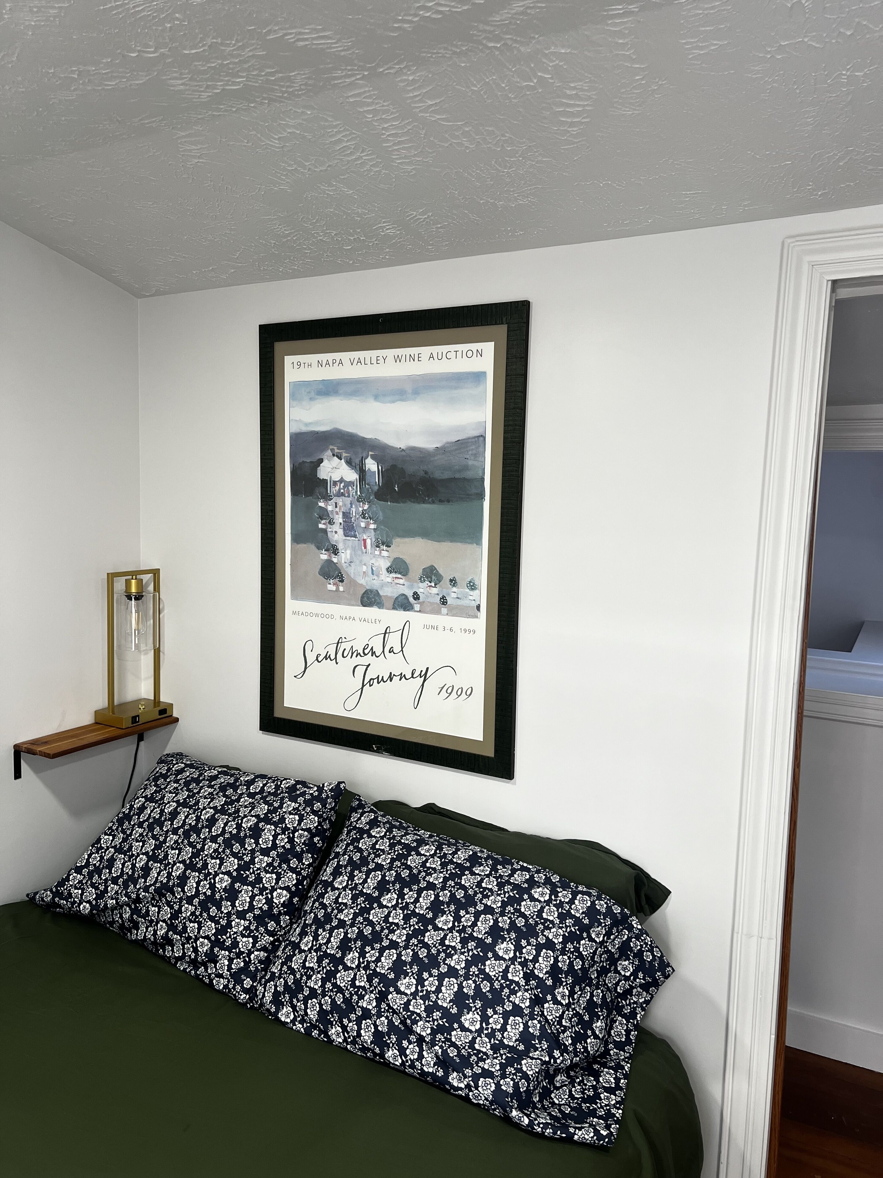 Ein Schlafzimmer mit einem Bett, einem gerahmten Bild an der Wand, einem Nachttisch mit Lampe und einer Tür zu einem weiteren Raum.