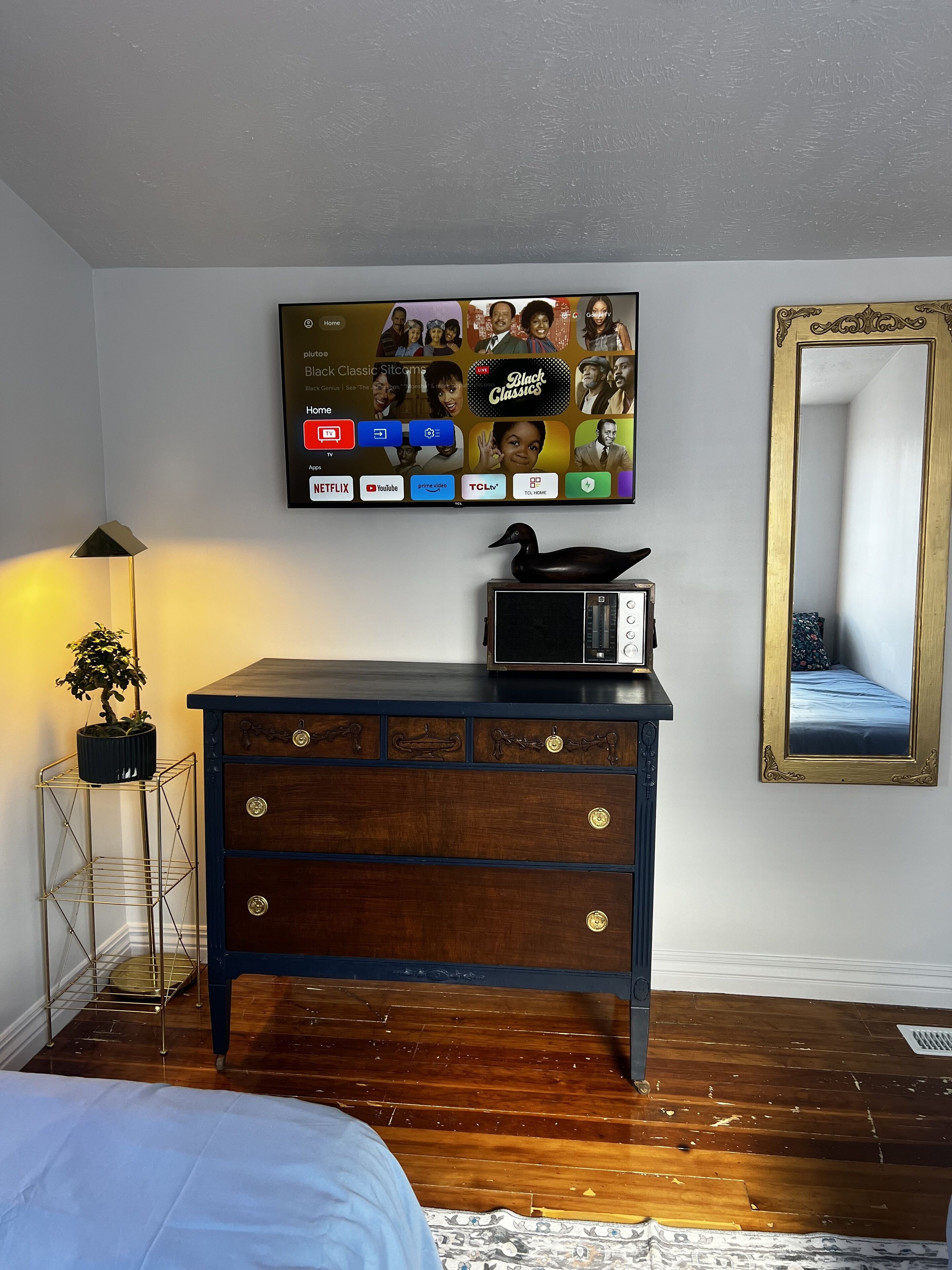 Un dormitorio con un aparador de madera, un televisor de pantalla plana montado en la pared mostrando la interfaz de un servicio de streaming, una radio pequeña y un espejo con un marco decorativo.