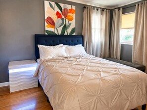 A bedroom with a bed, a nightstand, a wall painting, and a window with curtains.
