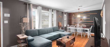 A modern living room with a sectional sofa, a dining table with chairs, a TV, and a wooden coffee table.