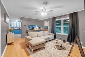 A living room with a sofa, a coffee table, a ceiling fan, and a view of the kitchen and outdoors through a window.