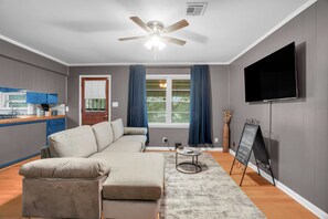 Un soggiorno moderno con un divano componibile, una TV a schermo piatto, una ventola a soffitto e un angolo cottura con mobili blu.