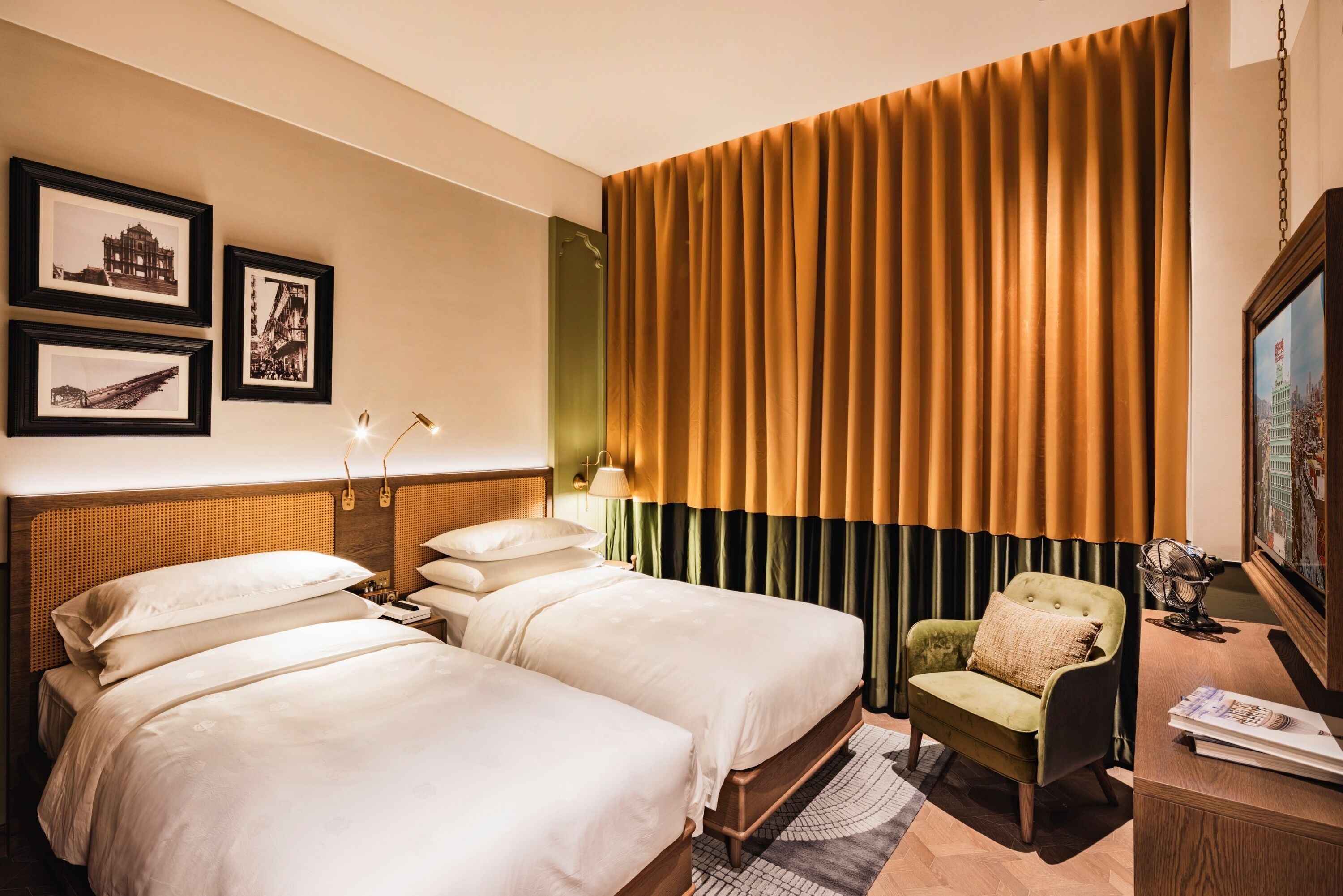 Une chambre d’hôtel avec deux lits, un fauteuil vert, un bureau avec une lampe et des tableaux encadrés au mur.