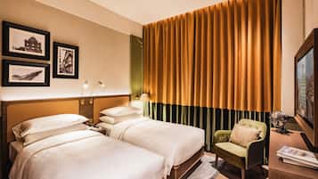 Une chambre d’hôtel avec deux lits, un fauteuil vert, un bureau avec une lampe et des tableaux encadrés au mur.