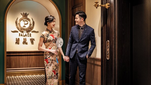A man and a woman holding hands in a hotel lobby with a sign reading "PALACE HOTEL" and a clock on the wall. - Hotel Central (Macau)