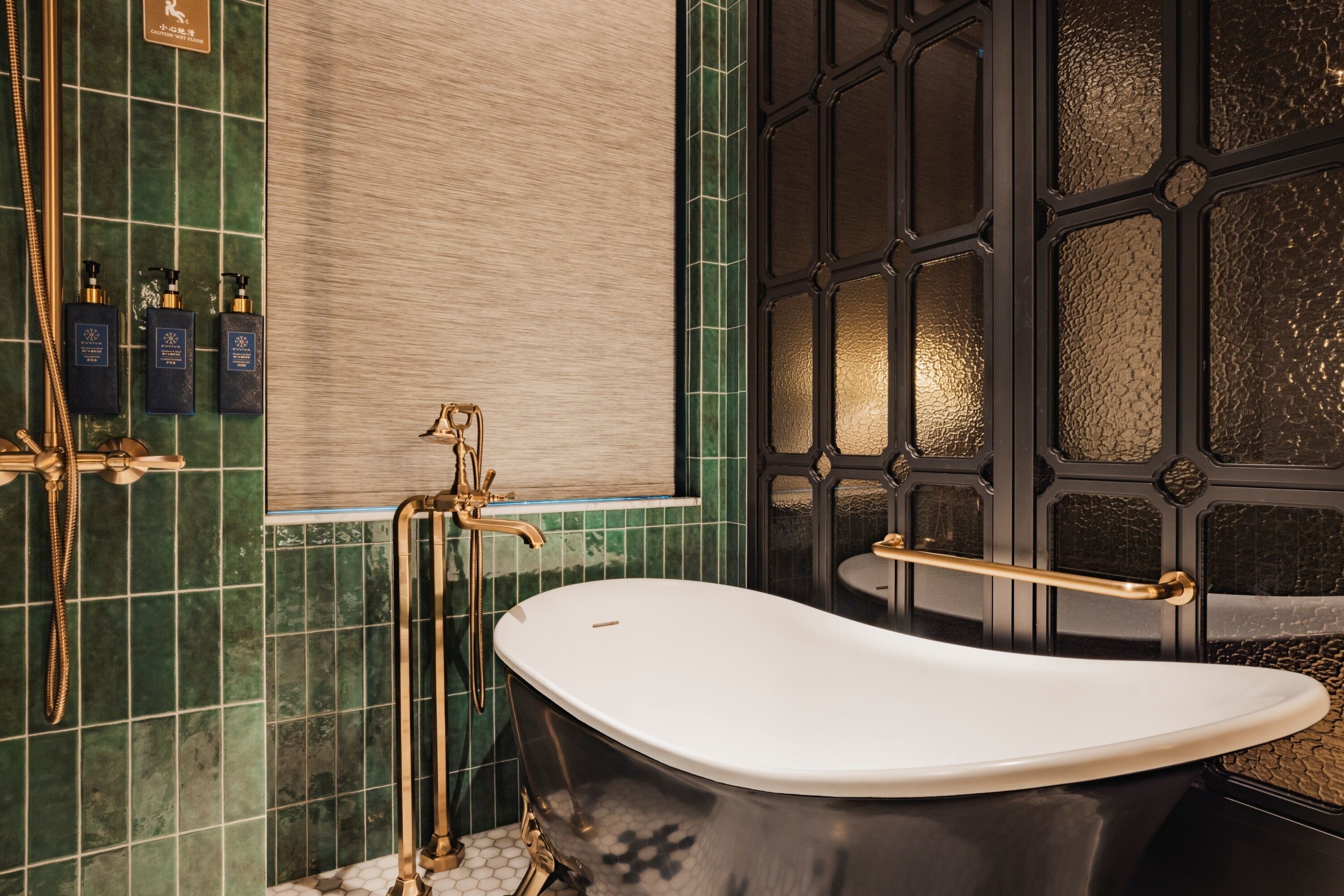 A bathroom with a freestanding bathtub, a shower with a gold faucet, and a tiled wall with a green pattern.