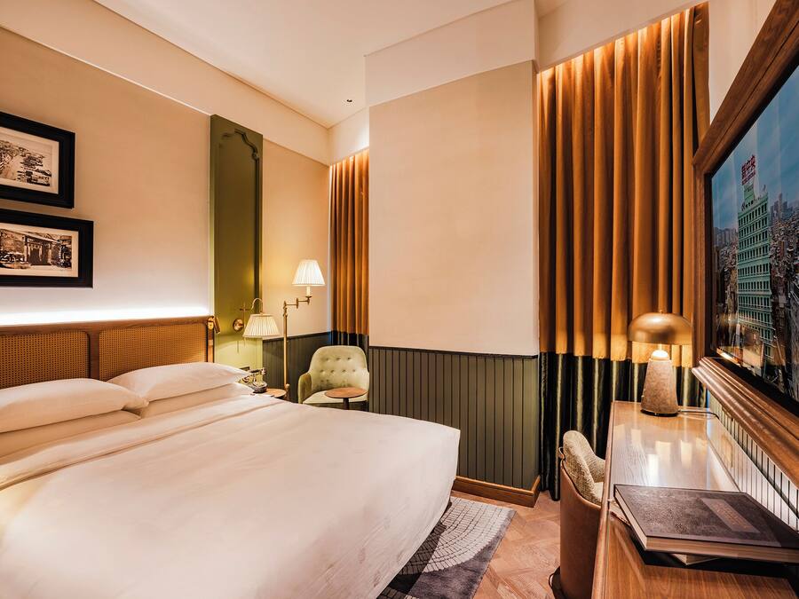 A hotel room with a large bed, a desk with a lamp, a chair, and a cityscape painting on the wall.