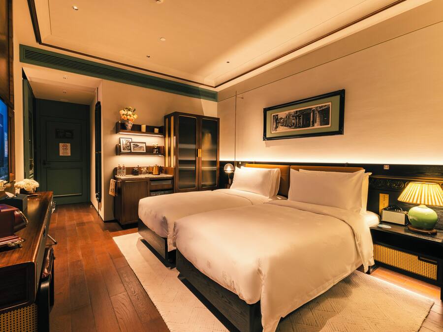 A hotel room with two beds, a desk, a wardrobe, and a nightstand with a lamp.
