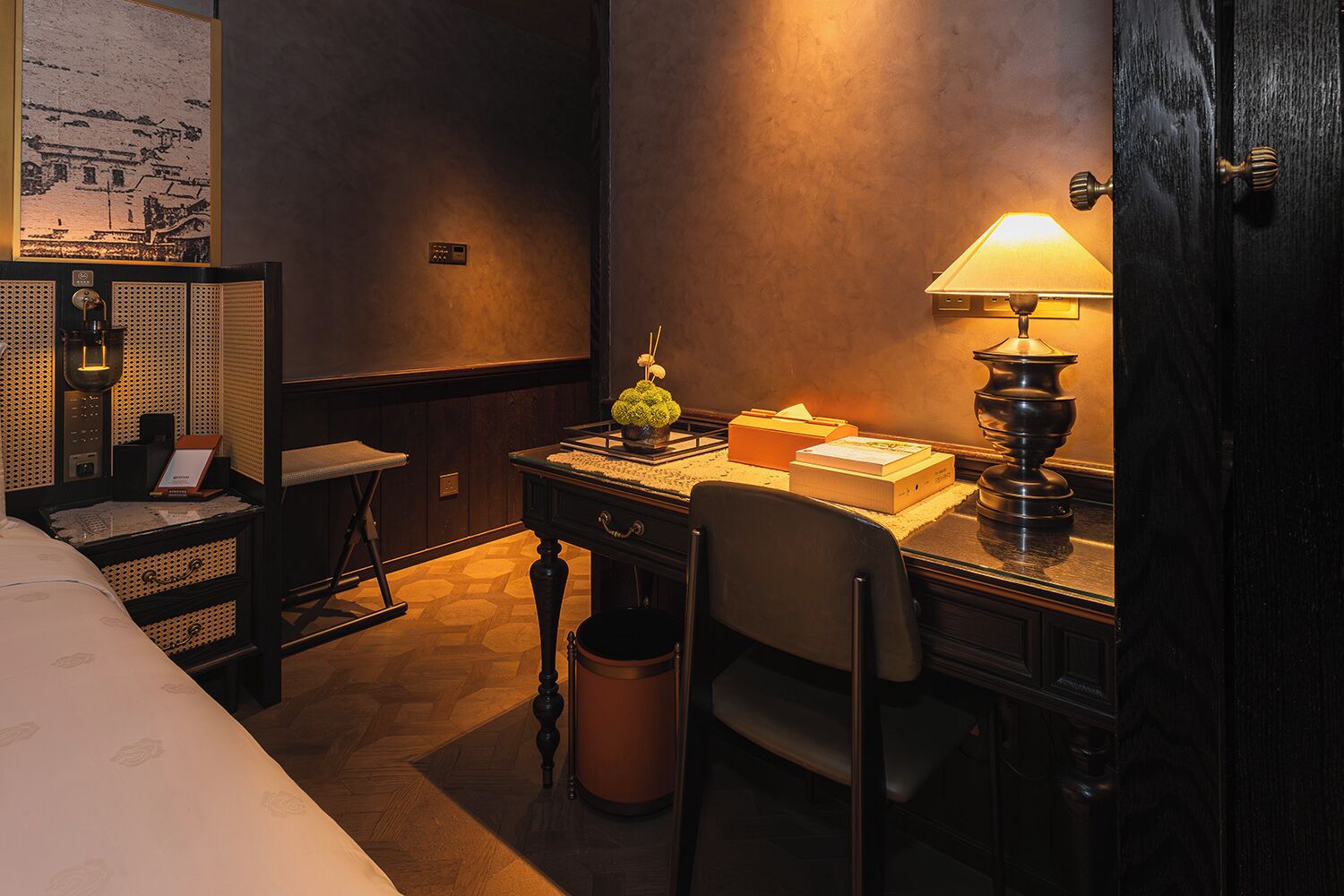 A hotel room with a bed, a desk with a lamp, a chair, a trash can, and a wall-mounted shelf with a plant.