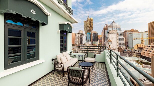 A balcony with a sofa, chairs, and a small table, overlooking a cityscape with various buildings. - Hotel Central (Macau)