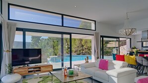 A modern living room with a large TV, white sofas, a coffee table, and a view of a swimming pool and greenery. - Villa Juno by Interhome (Jávea)