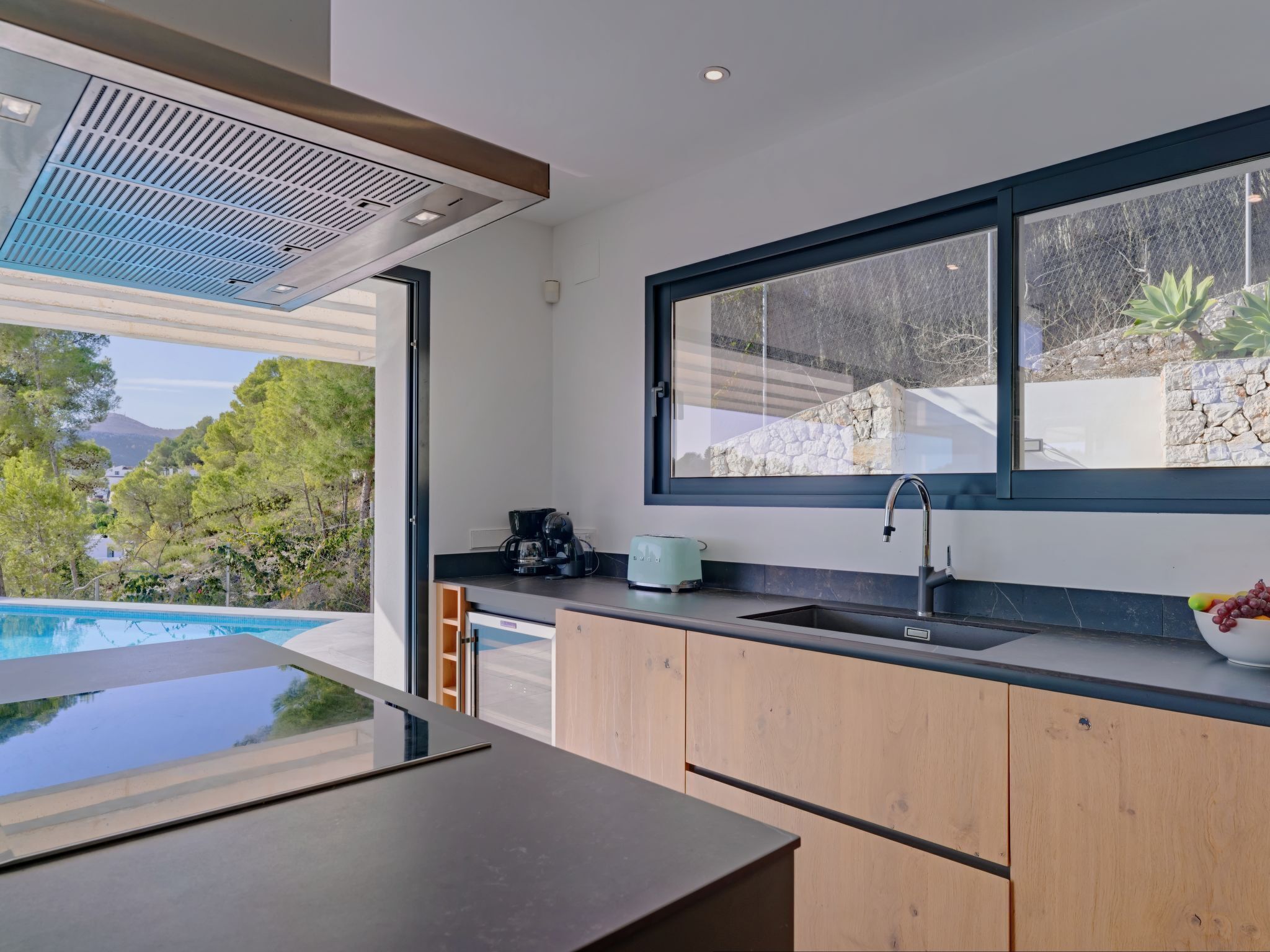 A modern kitchen with a large window, a built-in sink, and a view of a swimming pool and greenery.
