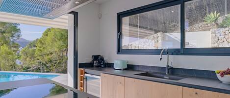 A modern kitchen with a large window, a built-in sink, and a view of a swimming pool and greenery.