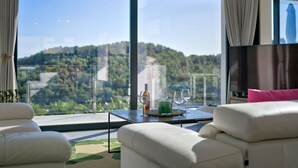 A modern living room with a large window offering a view of a forested hill. There is a white sofa, a small table with a glass of drink, and a flat-screen TV. - Villa Juno by Interhome (Jávea)