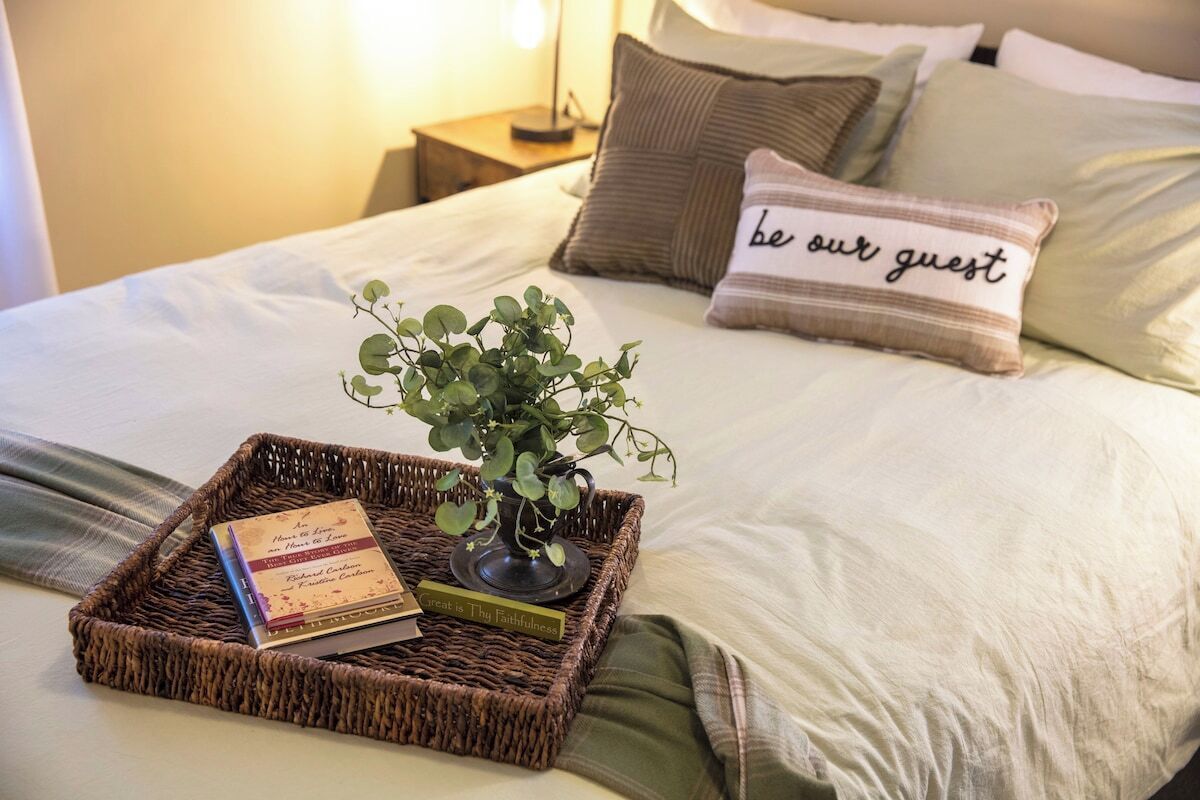 Een netjes opgemaakt bed met een sierkussen waarop staat 'wees onze gast', een kleine plant en een dienblad met een boek en een kaars.