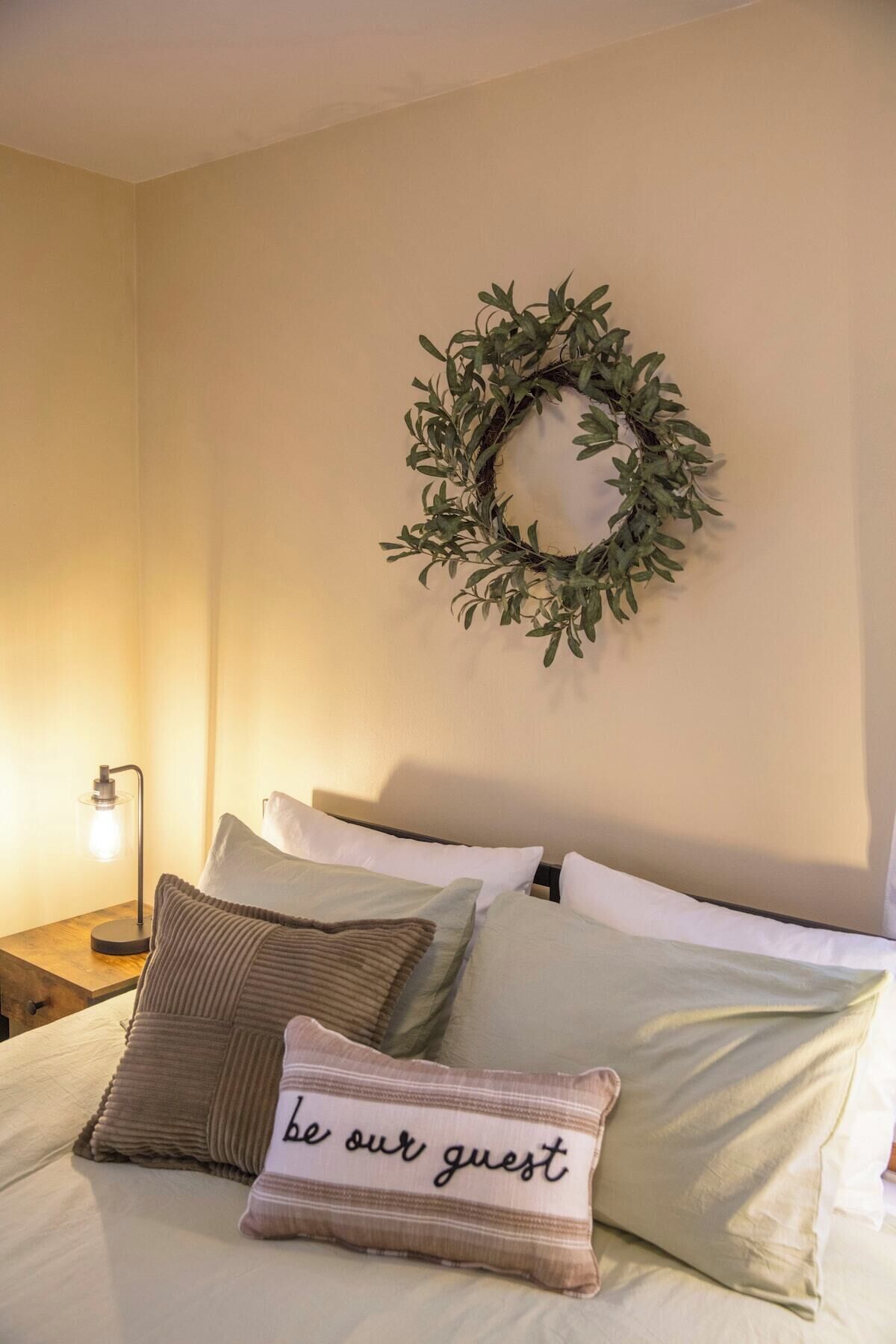 Un lit bien fait, avec un oreiller orné de l’inscription « soyez les bienvenus », une couronne décorative accrochée au mur et une lampe de chevet.