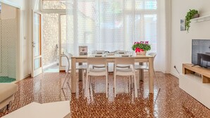 Un comedor con una mesa preparada para cuatro personas, un televisor sobre un mueble bajo y una planta en el alféizar de la ventana.