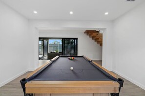 A pool table in a room with white walls, a wooden staircase, and a view through a glass door.