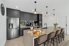 A modern kitchen with a stainless steel refrigerator, a large island with bar stools, pendant lighting, and a built-in oven.