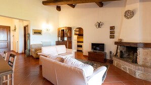 Un salón amplio con un sofá blanco, vigas de madera, una chimenea y una zona de cocina al fondo.