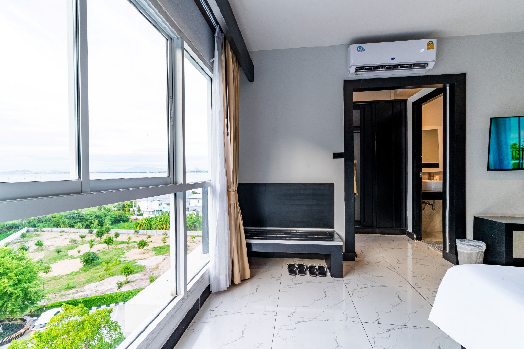 A hotel room with a large window, a bed, a bench, and a view of a green landscape.
