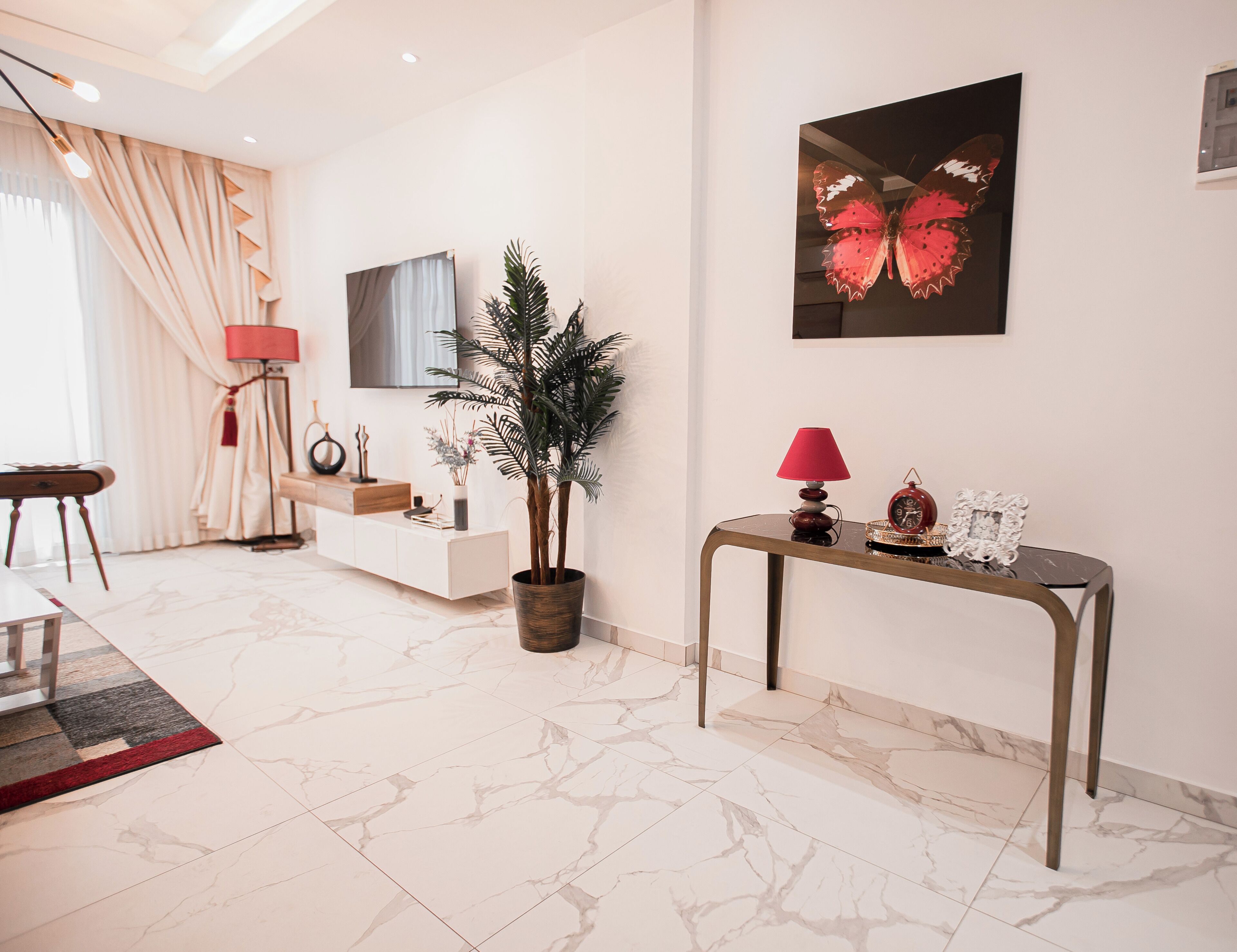 A modern living room with a marble floor, a white console table, a red lamp, a potted plant, and a wall-mounted TV.