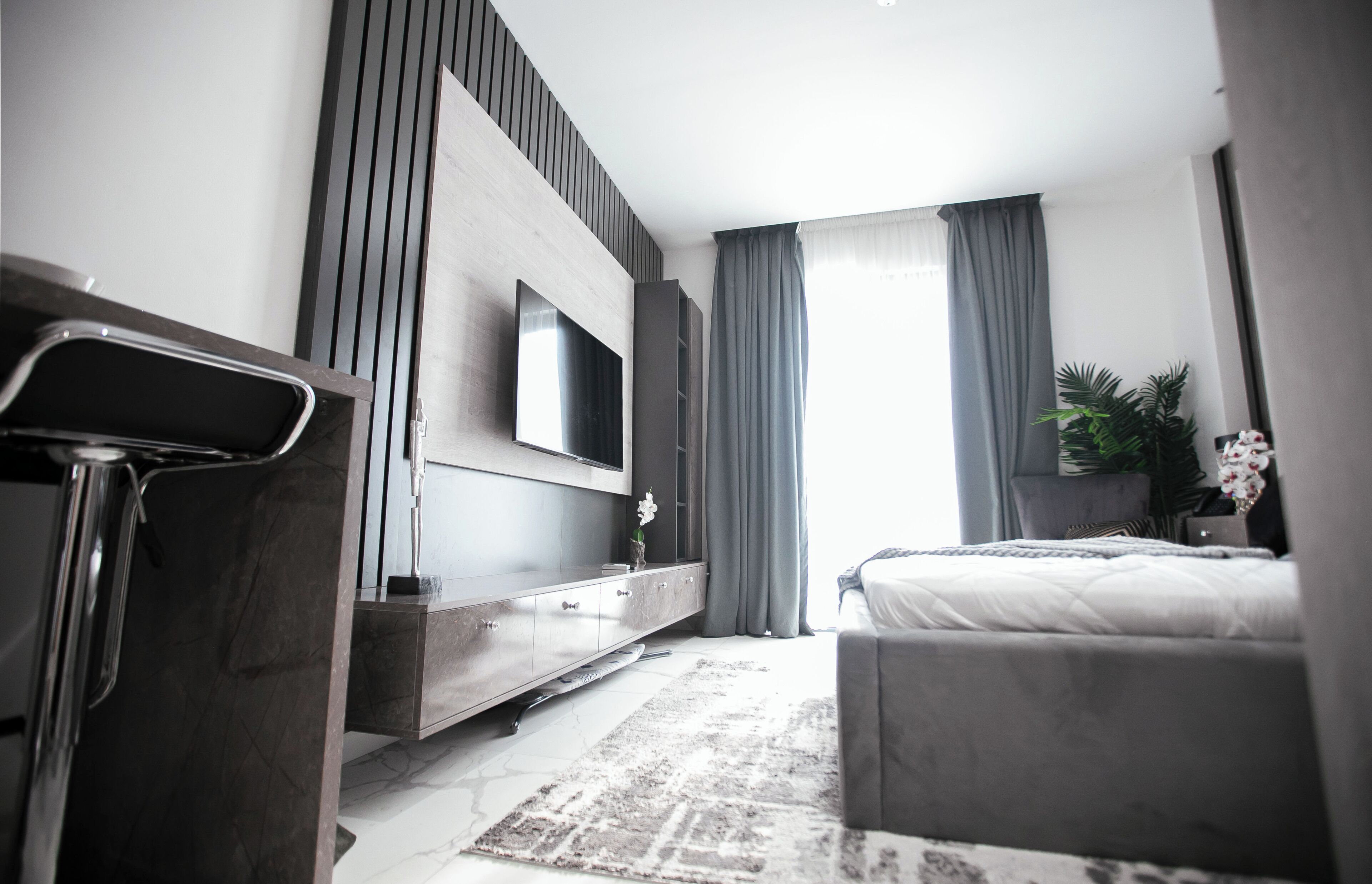 A modern hotel room with a bed, a TV mounted on the wall, a bedside table, and a large window with curtains.