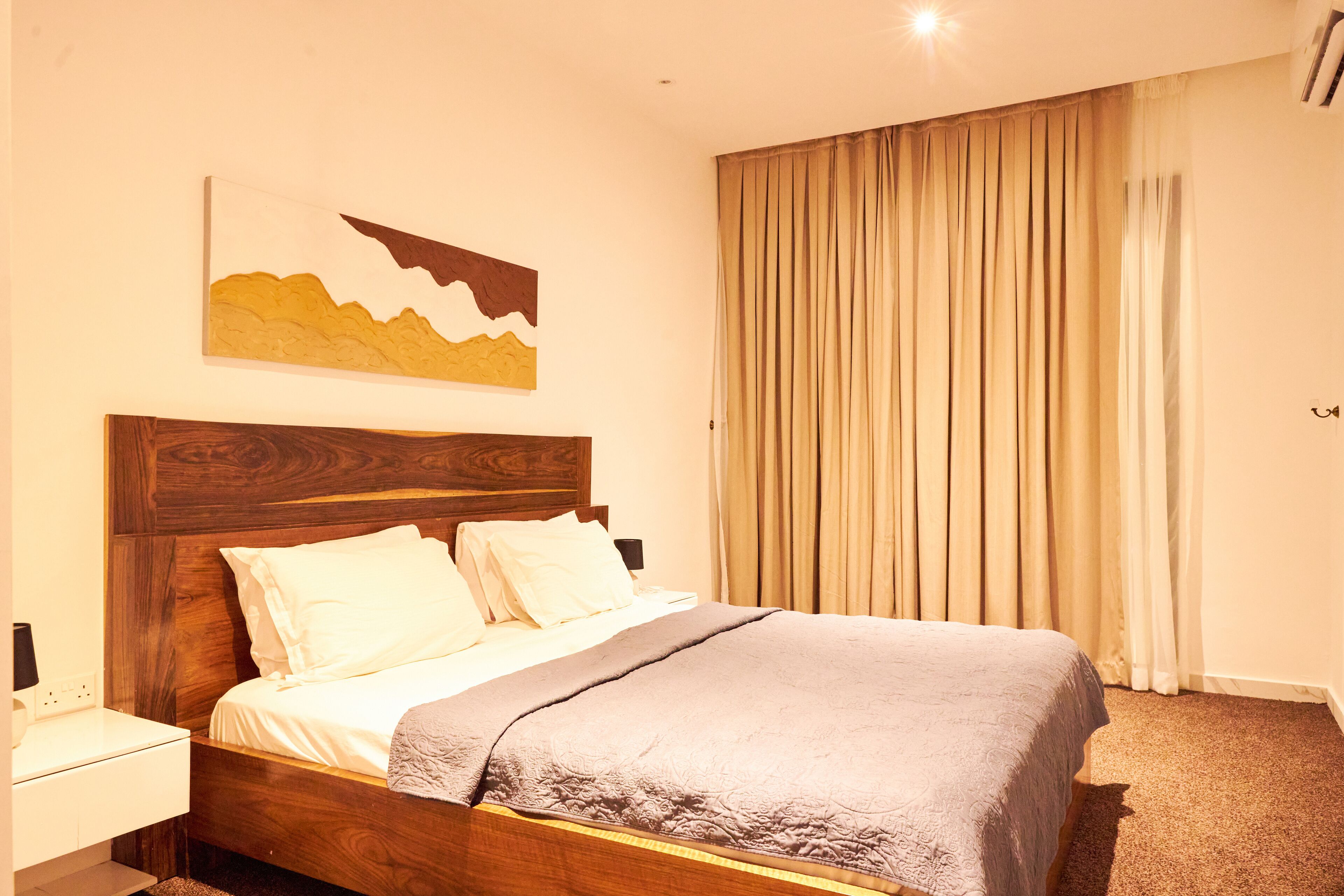 A neatly made bed with a wooden headboard, a bedside table, and a large window with curtains.