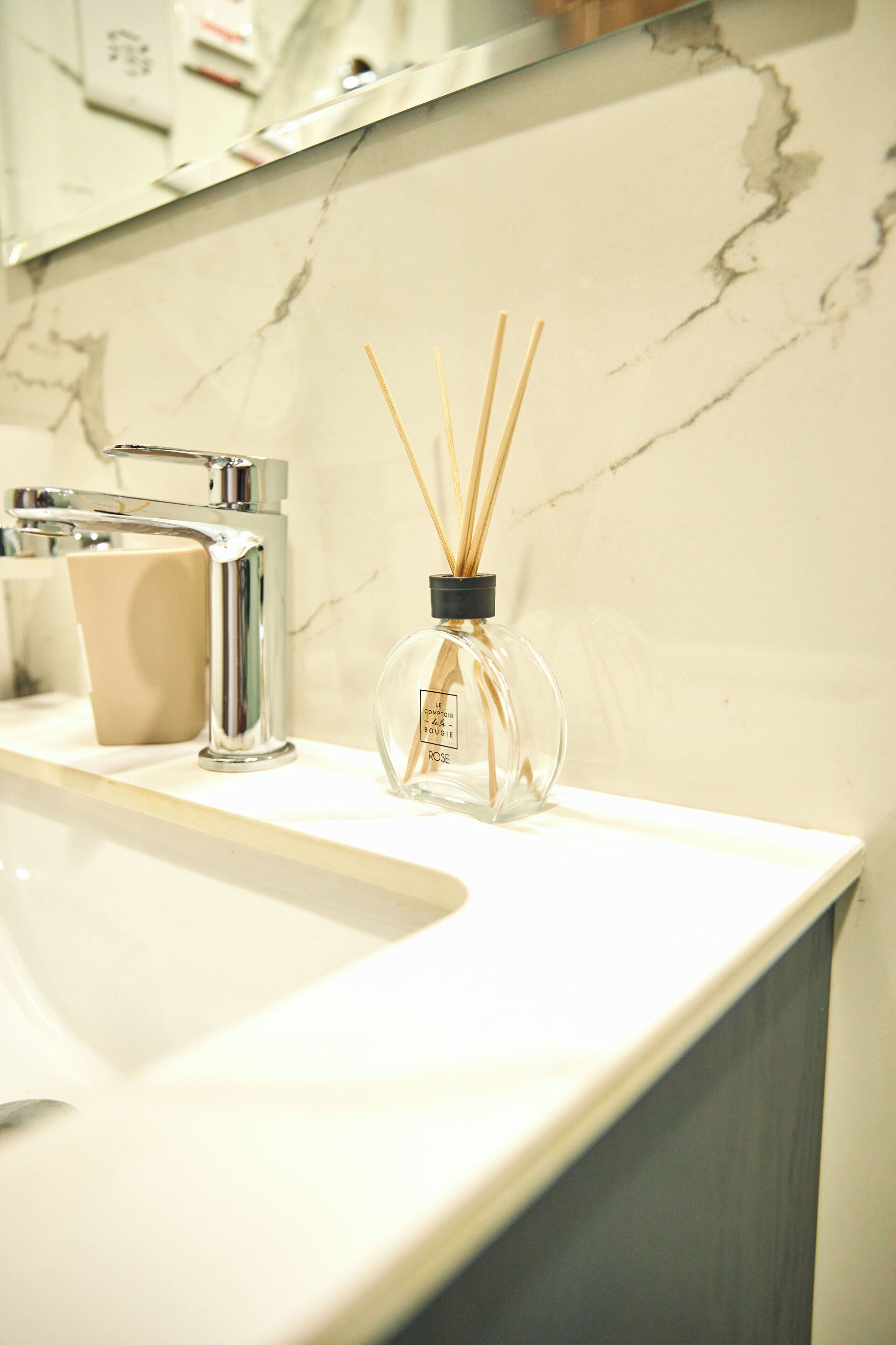A bathroom sink with a marble countertop, a modern faucet, a beige soap dispenser, and a glass bottle with reed sticks.