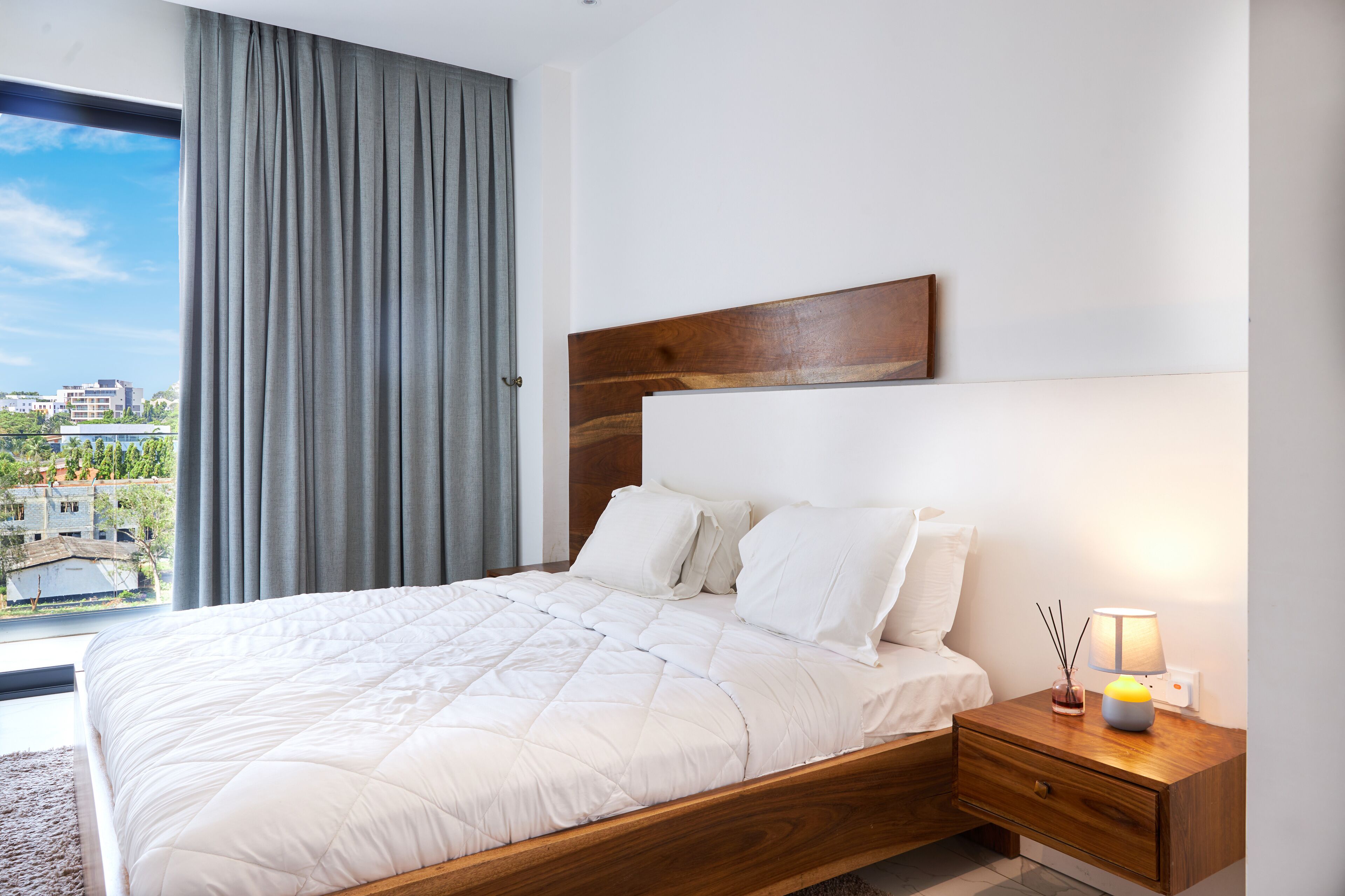 Un dormitorio con una cama grande, cabecera de madera, mesita de noche con lámpara y vista a edificios por la ventana.