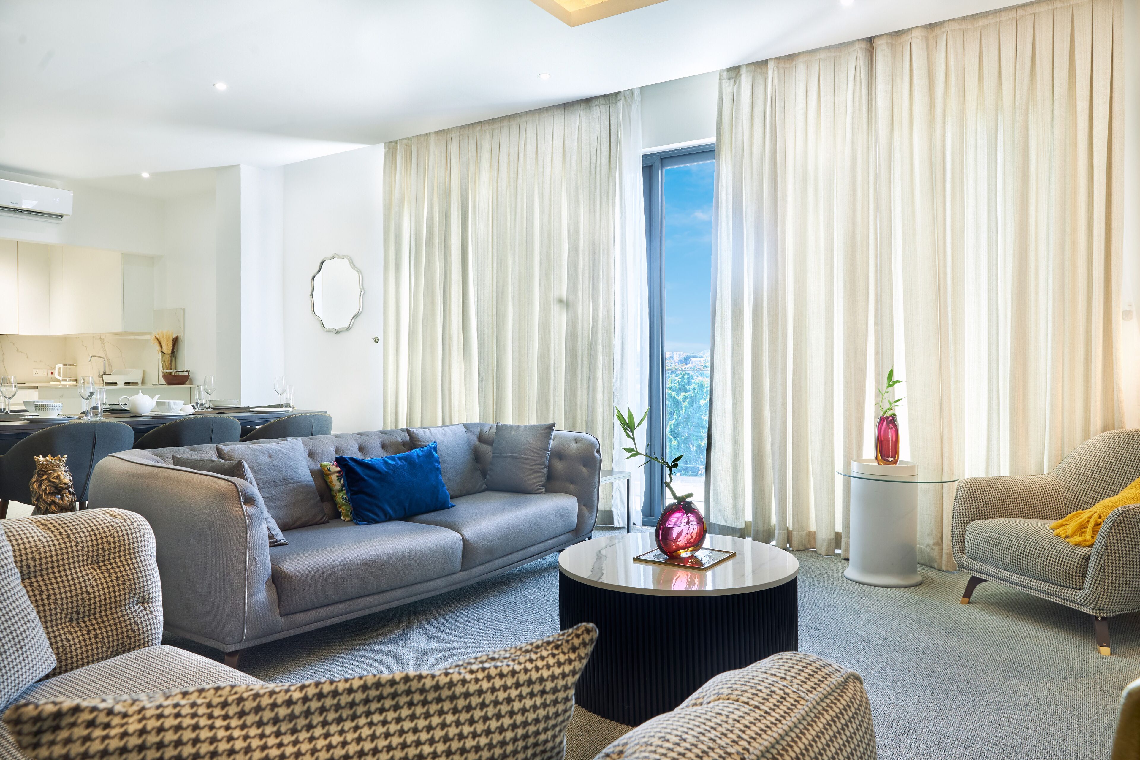 A modern living room with a grey sofa, patterned armchairs, a round coffee table, and a view of the city through large windows.
