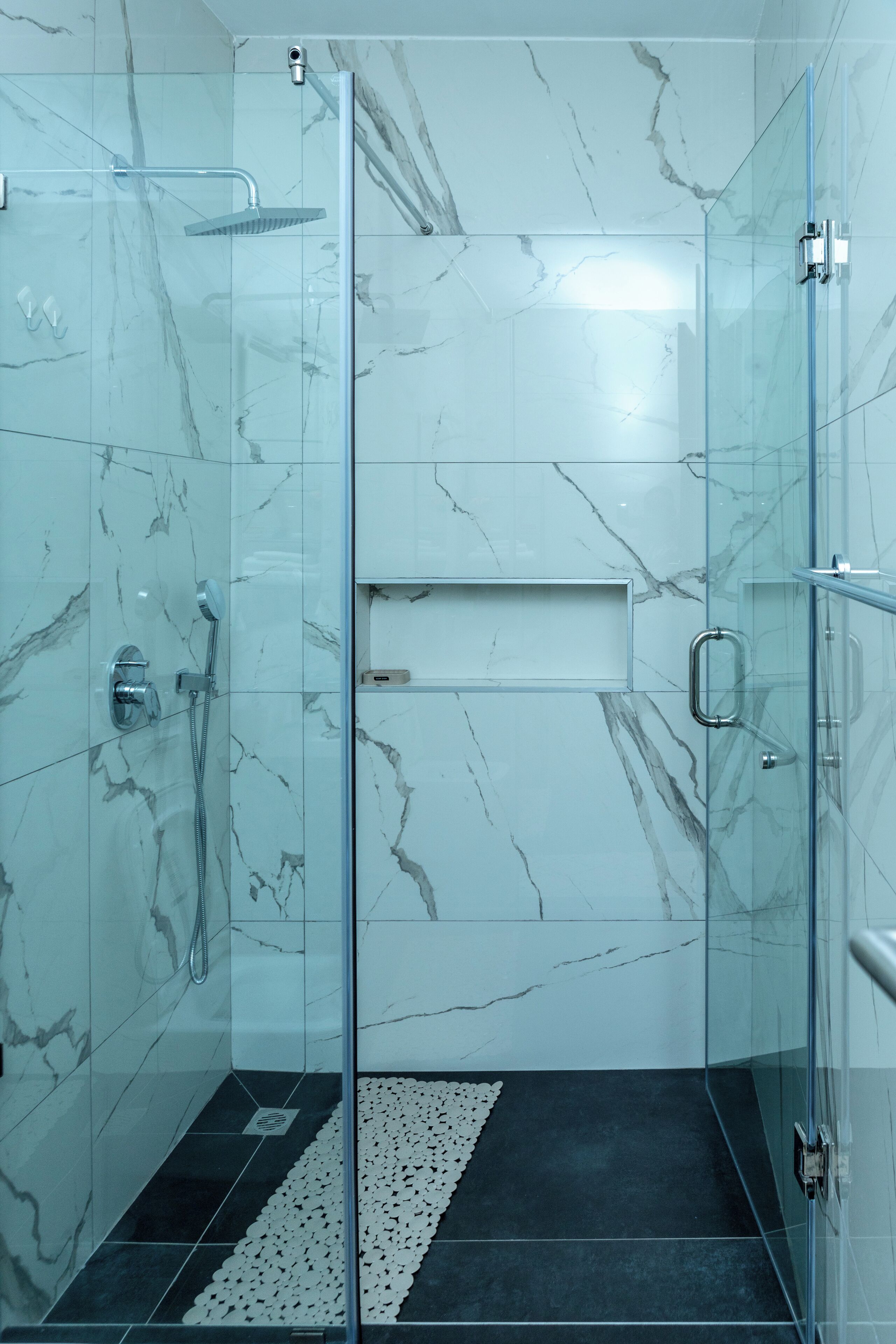 Une douche fermée par des parois de verre, avec un mur en marbre, un tapis de sol noir et une petite étagère.