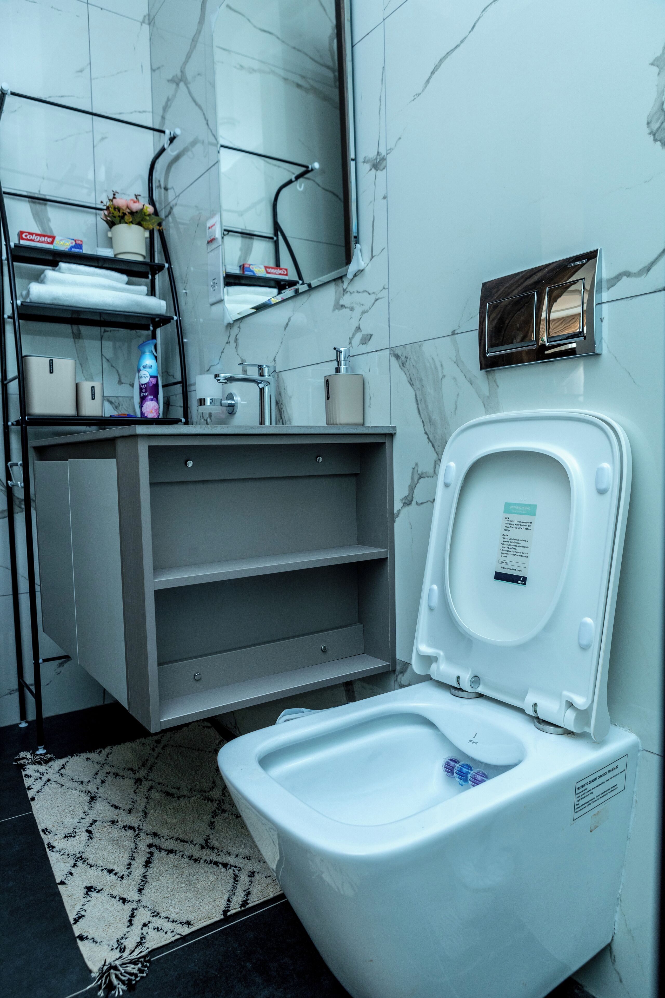 A modern bathroom with a marble wall, a toilet, a sink with a mirror, and a shelf with toiletries.