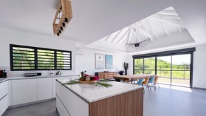 Una cocina moderna con una isla central, un comedor con una mesa grande y vistas a zonas verdes a través de las ventanas.