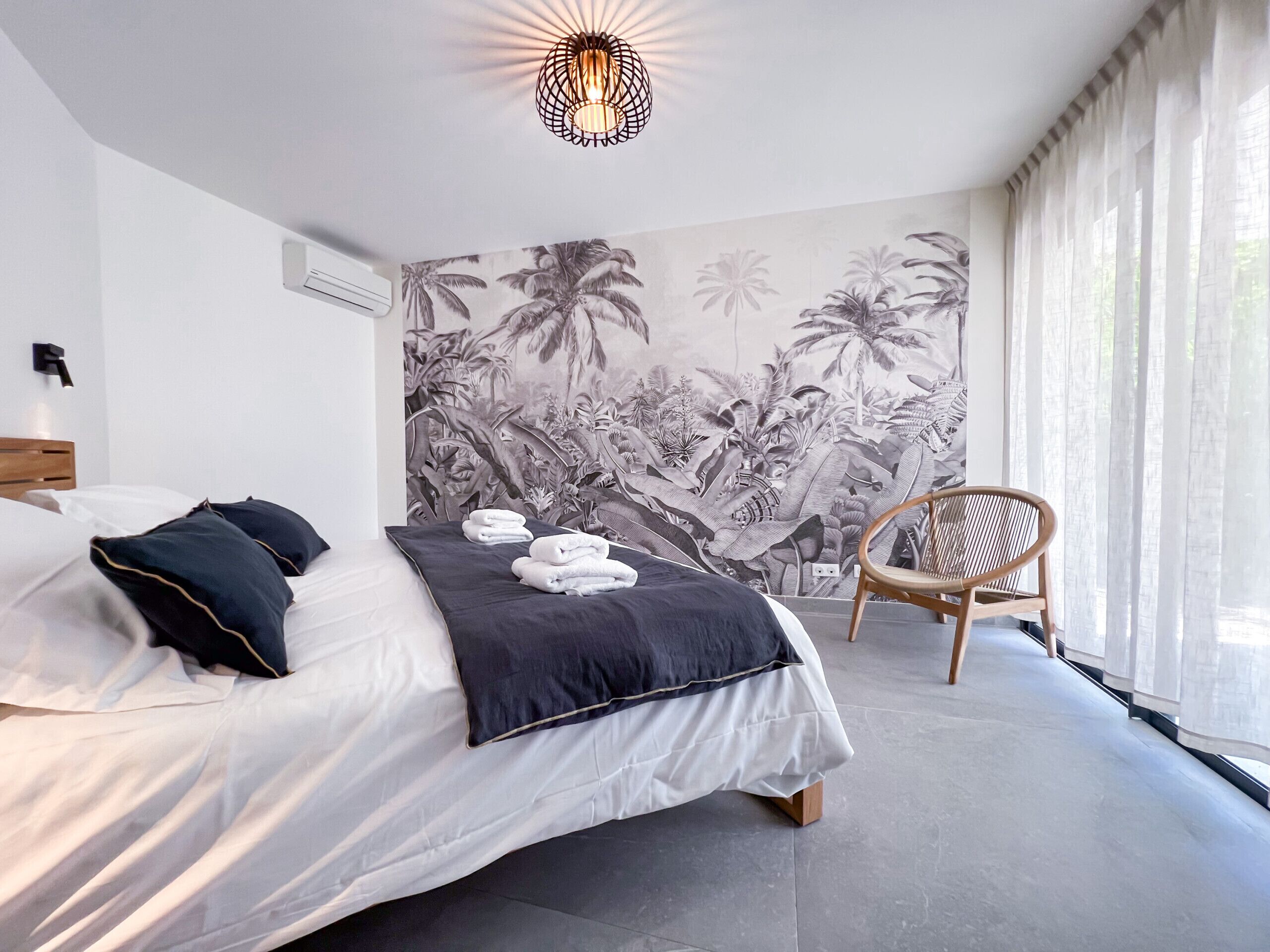 A modern bedroom with a bed, a chair, and a tropical mural on the wall.