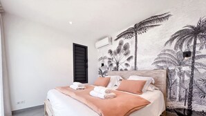 Un dormitorio con cama, almohadas, una colcha, una mesita de noche y una pared con un mural tropical.