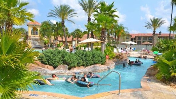 Zona de piscina con gente dentro de flotadores, palmeras y un edificio al fondo.