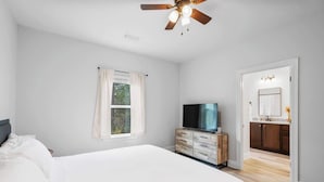 Ein Schlafzimmer mit Bett, Fernseher, einem hölzernen Schminktisch und Deckenventilator. Ein Badezimmer ist durch eine geöffnete Tür sichtbar.