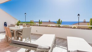 En balkon med et bord og stole, en liggestol og udsigt til havet og bygninger.