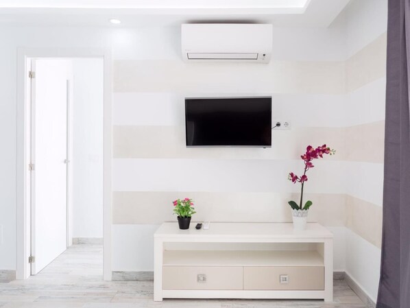 A modern hotel room with a flat-screen TV mounted on the wall, a white console table with two potted plants, and an air conditioning unit above the TV. - 1713 Sunset ,ocean view terrace,heated pool (Costa Adeje)