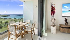 A balcony with a table and chairs, a large window with a view of the ocean, and a mounted artwork. - b703 (Cupecoy)