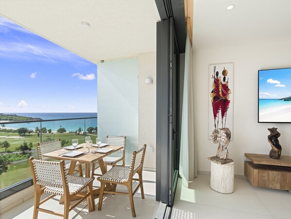 A balcony with a table and chairs, a large window with a view of the ocean, and a mounted artwork.